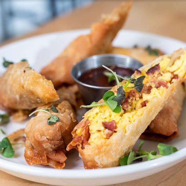 Golden-fried breakfast egg rolls filled with scrambled eggs and bacon, garnished with microgreens and dipping sauce.