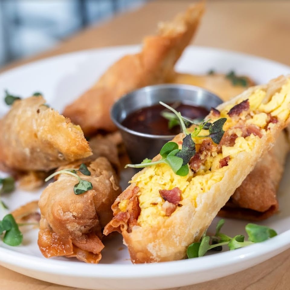 Golden-fried breakfast egg rolls filled with scrambled eggs and bacon, garnished with microgreens and dipping sauce.