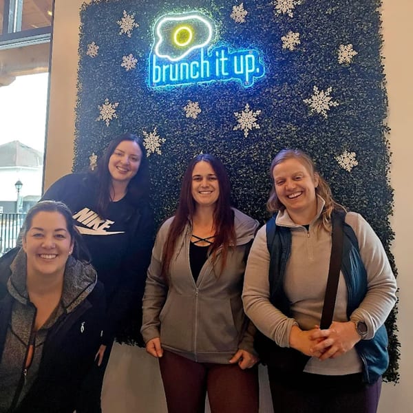 Four smiling women posing in front of a green wall with a neon "brunch it up" sign.