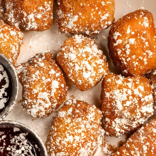 Close-up of golden-brown beignets dusted with powdered sugar, with two small cups of chocolate sauce.
