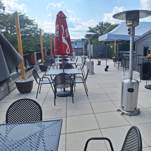 Outdoor patio with black metal tables and chairs, red umbrellas, and patio heaters under a cloudy sky.