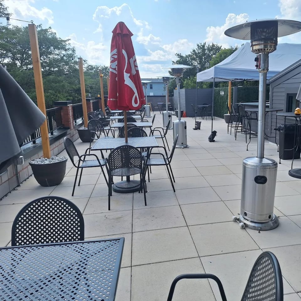 Outdoor patio with black metal tables and chairs, red umbrellas, and patio heaters under a cloudy sky.