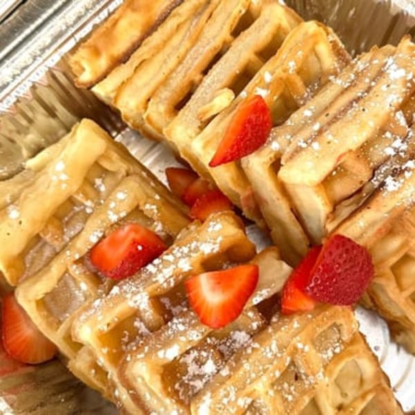 Golden waffles topped with powdered sugar and fresh strawberry slices, served in a foil container.