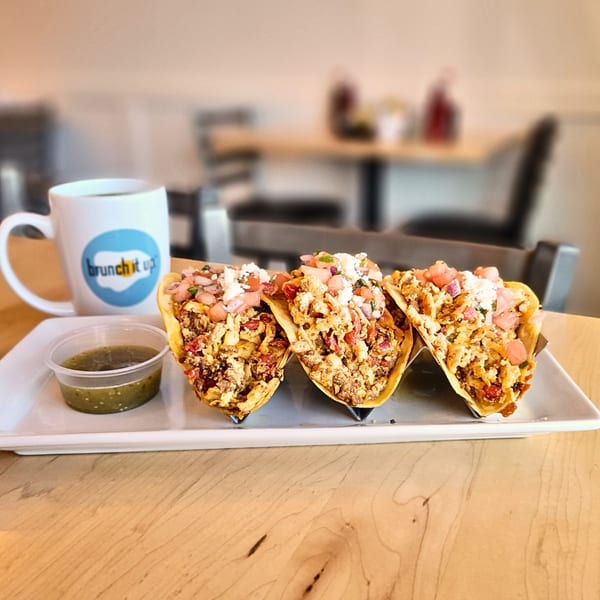 Three breakfast tacos with scrambled eggs, salsa, and cheese, alongside a coffee mug and green salsa.