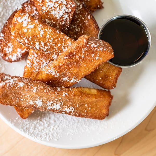 Golden-brown French toast sticks, dusted with powdered sugar, served with a side of syrup.