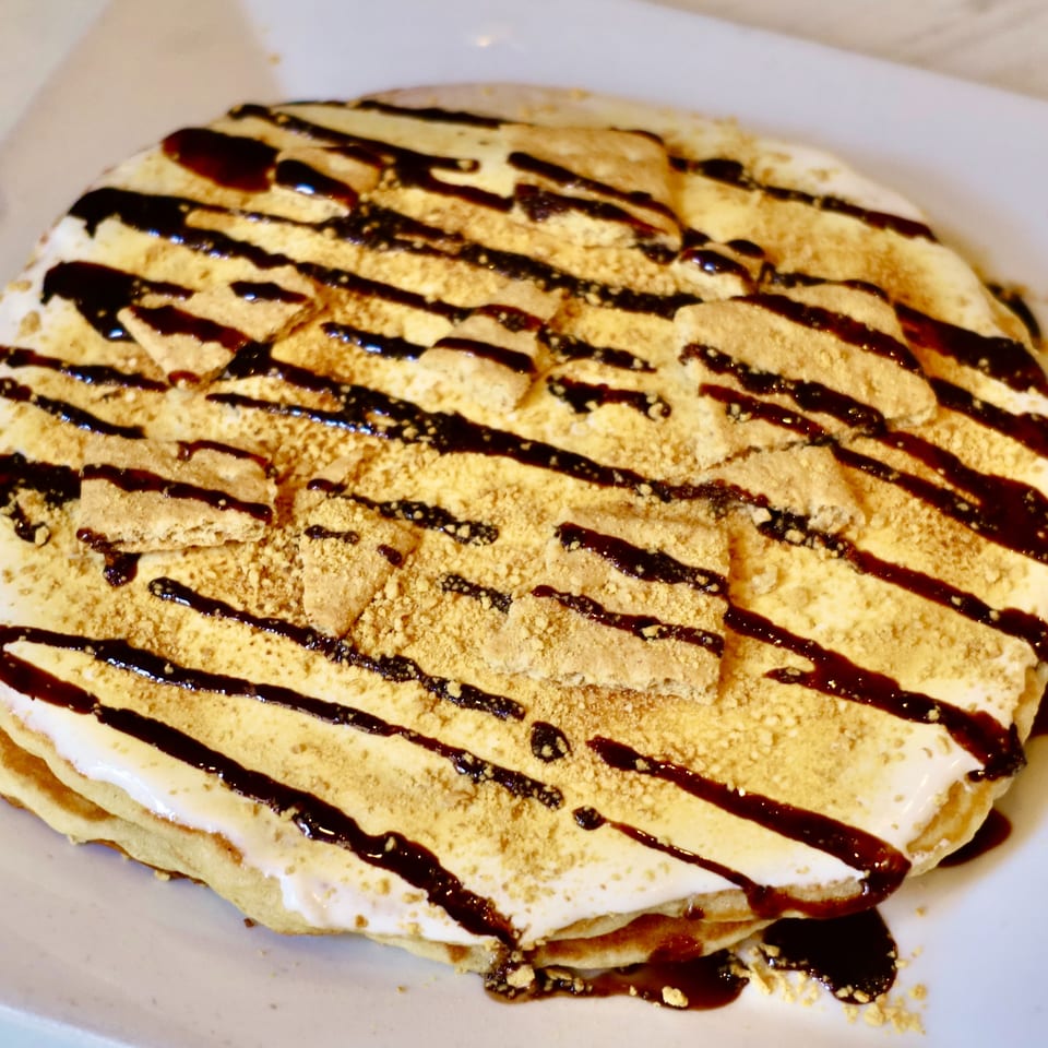 A stack of pancakes topped with whipped cream, chocolate syrup, and graham cracker crumbles.