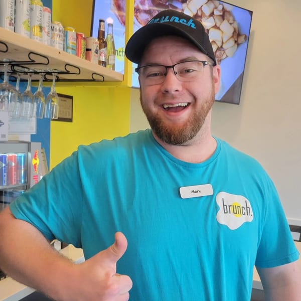 Smiling male server in a "Brunch" hat and shirt gives a thumbs up.
