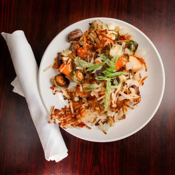 A white plate of crispy hash browns, roasted vegetables, and green onions on a dark wooden table.