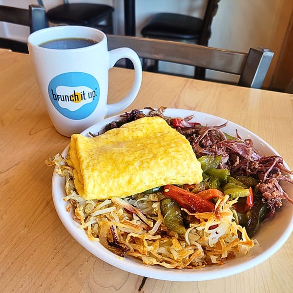 A Brunch It Up mug of coffee next to a plate of omelet, hash browns, and shredded meat.