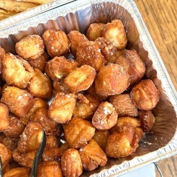 A foil tray filled with golden-brown, powdered sugar-dusted beignets, ready for a brunch service.