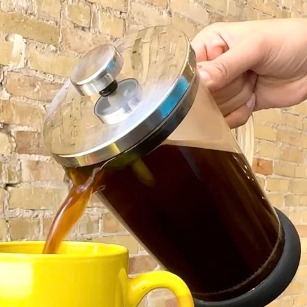 A hand pours dark coffee from a French press into a bright yellow mug against a brick wall.
