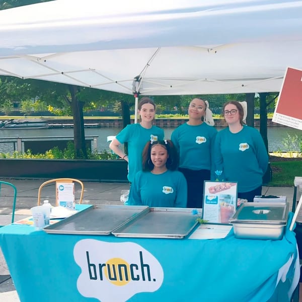 Four women in "Brunch." branded shirts stand behind a table with food trays and signage.