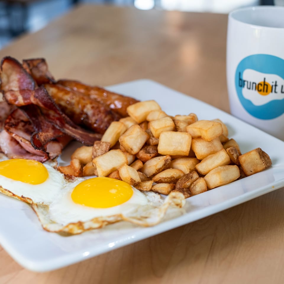 A Brunch It Up breakfast plate with fried eggs, bacon, ham, sausages, and home fries.