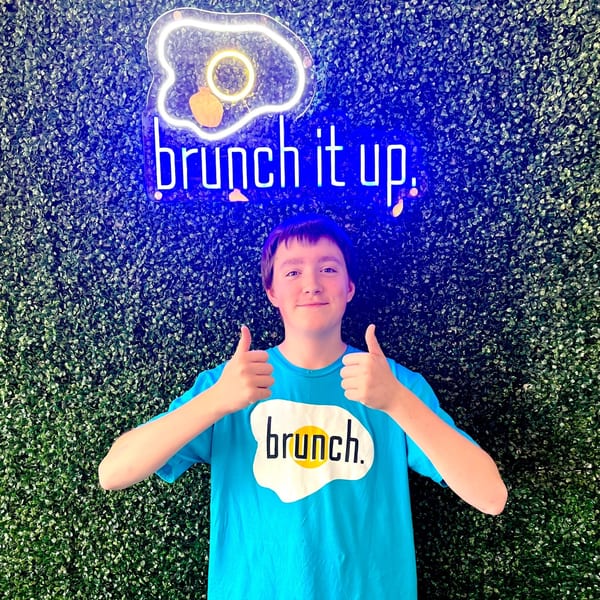 A person in a blue "Brunch" t-shirt gives two thumbs up in front of a neon "brunch it up" sign.