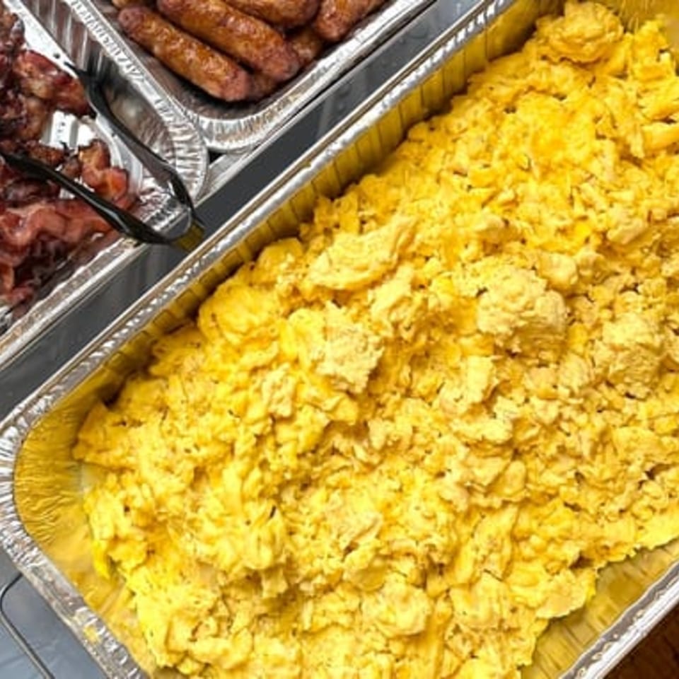 Scrambled eggs in a foil pan, with sausages and bacon in another pan in the background.