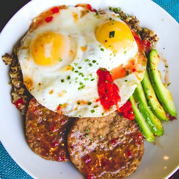 A brunch plate with two fried eggs, sausage patties, avocado slices, and quinoa, drizzled with sauce.
