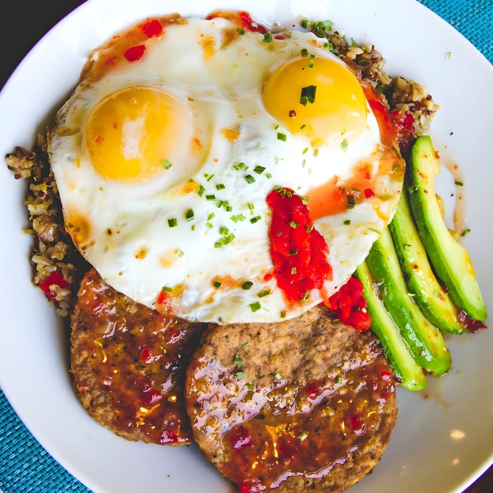 A brunch plate with two fried eggs, sausage patties, avocado slices, and quinoa, drizzled with sauce.